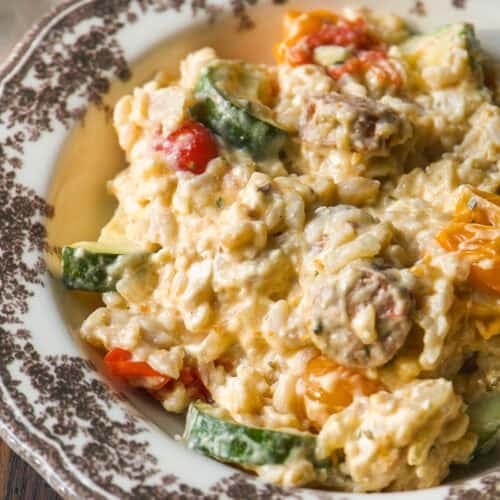 Creamy rice dish with zucchini, cherry tomatoes, and mushrooms, served on a vintage floral-patterned plate atop a rustic wooden table.