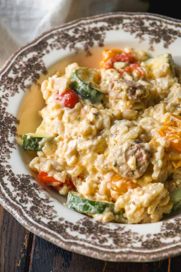Creamy rice dish with zucchini, cherry tomatoes, and mushrooms, served on a vintage floral-patterned plate atop a rustic wooden table.