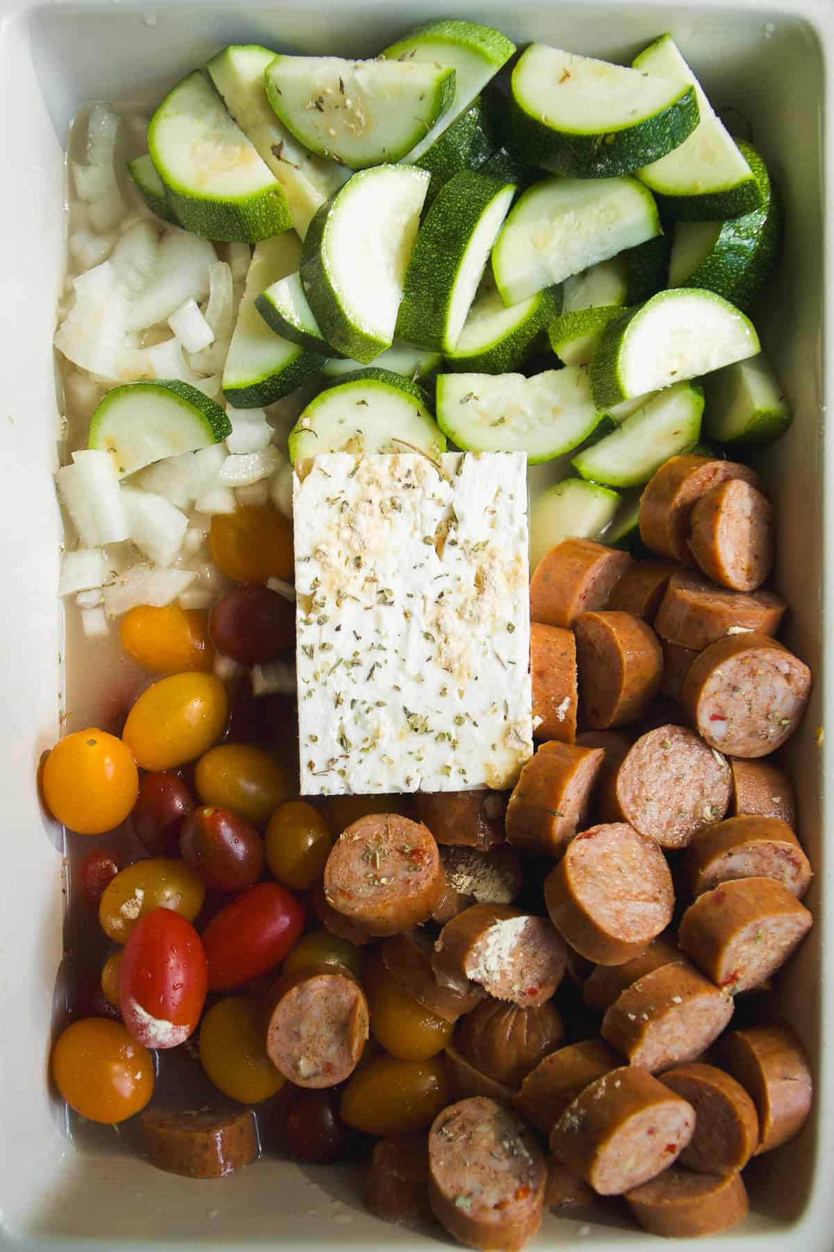 Sliced zucchini, diced onion, a block of seasoned feta, halved cherry tomatoes, and sliced sausage are arranged in sections in a white baking dish, ready for roasting.