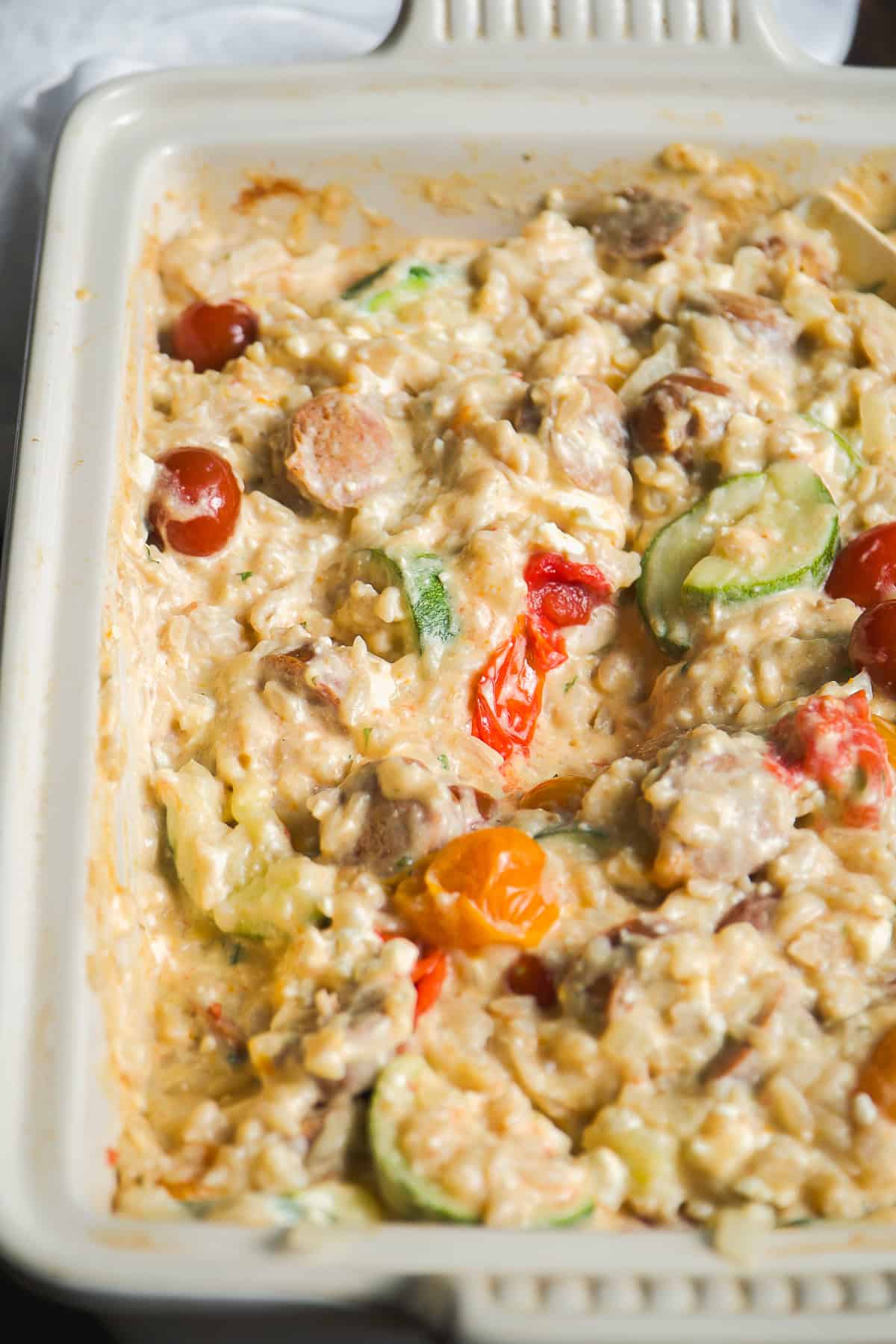 A close-up of a creamy casserole in a white baking dish with visible zucchini slices, cherry tomatoes, sausage pieces, and melted cheese.