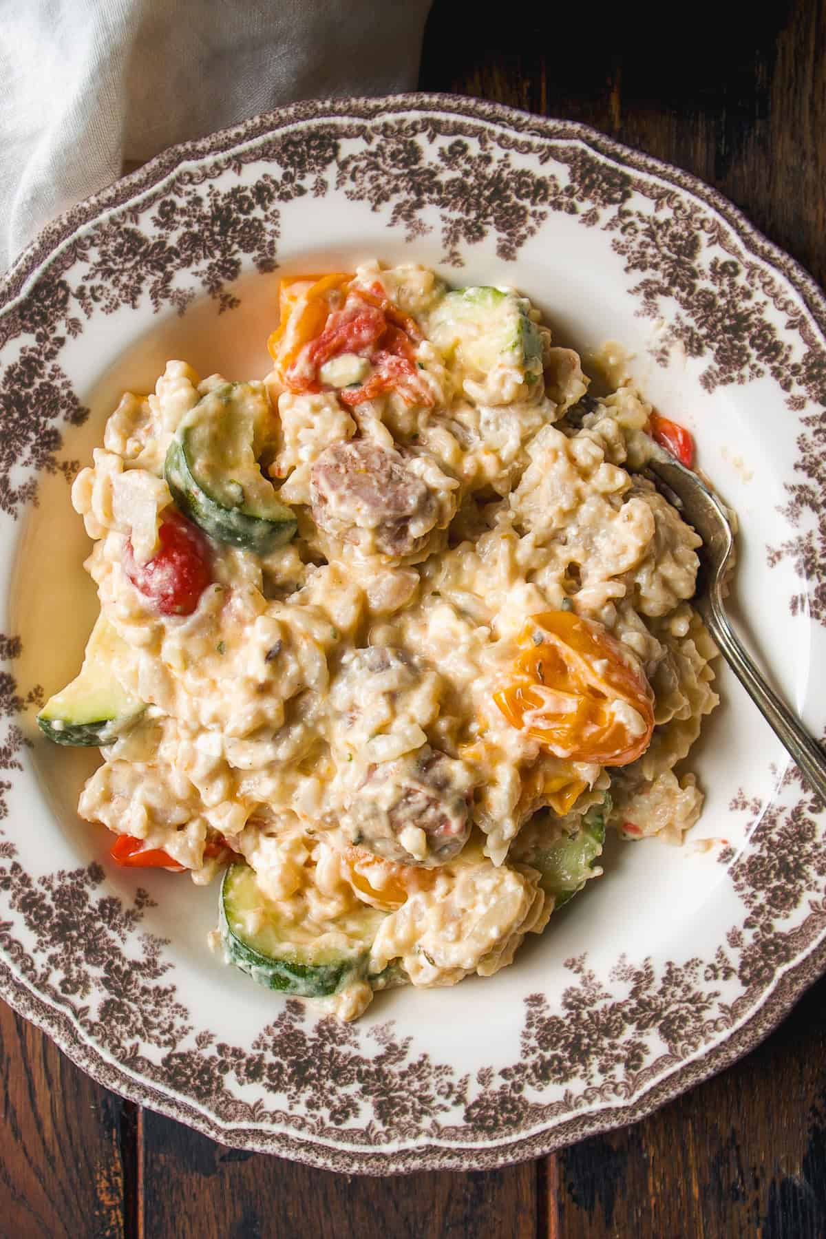 A vintage plate with creamy pasta, sausage slices, zucchini, and cherry tomatoes in a rich sauce, placed on a dark wooden table with a silver fork.