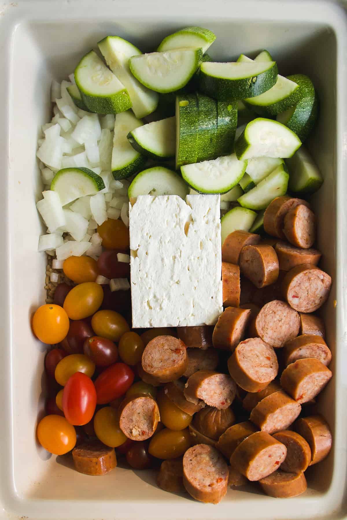 Chopped zucchini, onions, grape tomatoes, sausage slices, and a block of feta cheese arranged in sections in a white baking dish.