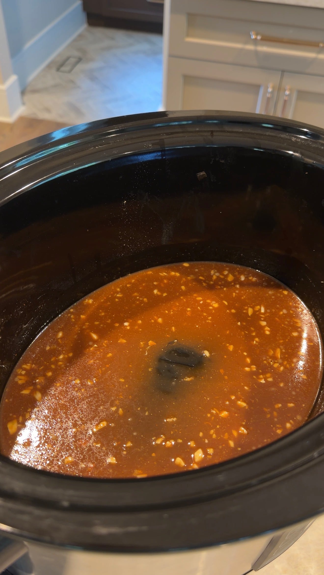 A close-up of a slow cooker filled with a brown, chunky liquid sauce or broth, possibly with garlic or onions, sitting on a kitchen countertop.