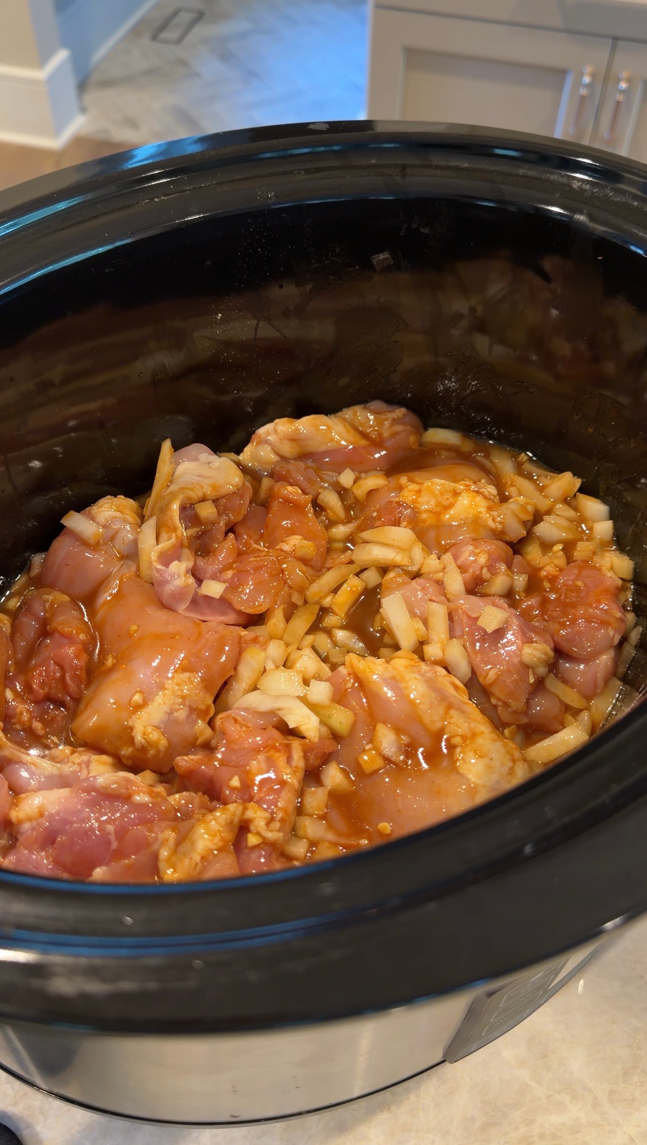 Raw chicken pieces mixed with chopped onions and sauce inside a black slow cooker, ready to be cooked.