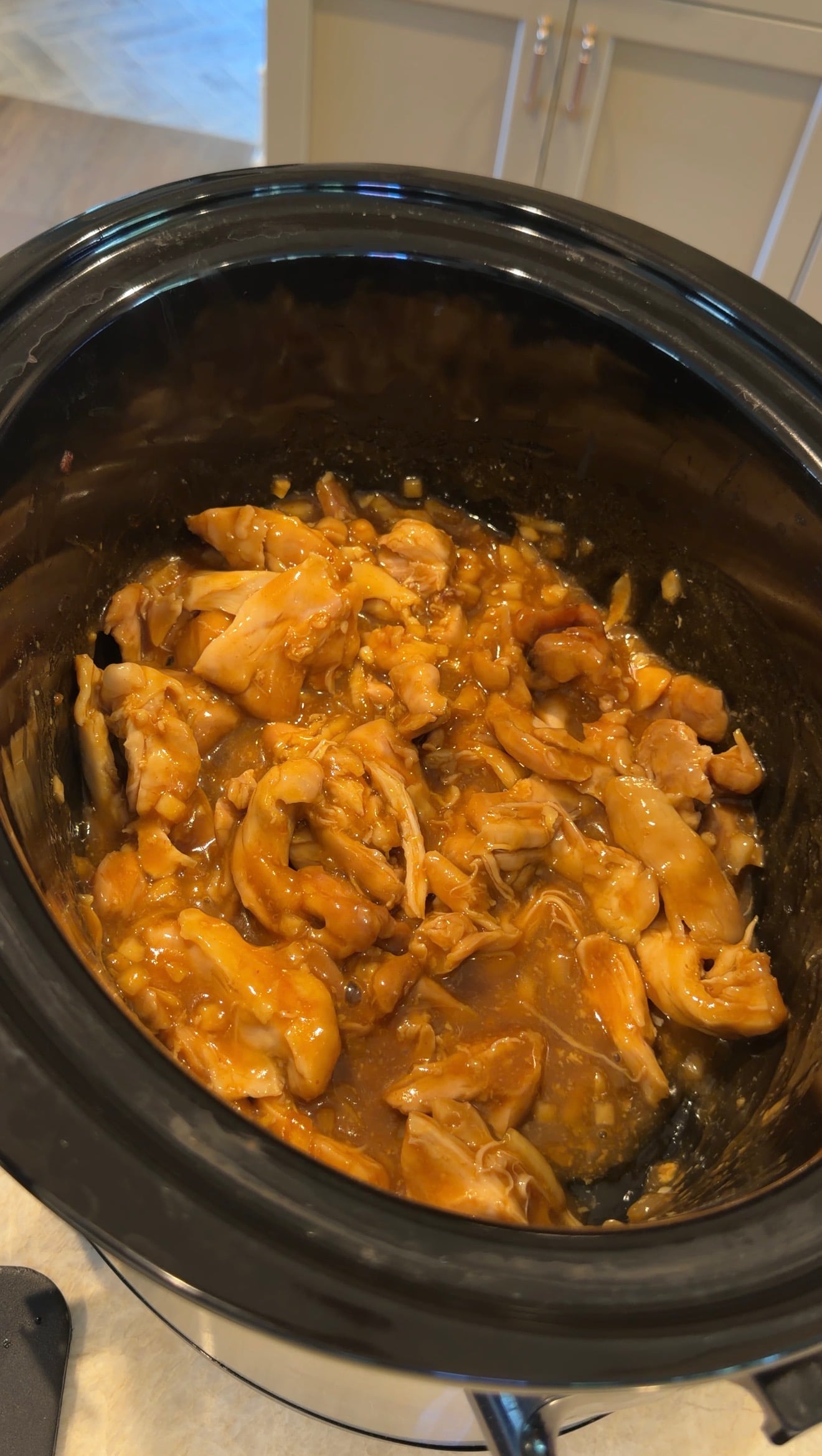 Shredded chicken in a thick brown sauce cooking inside a black slow cooker, with kitchen counters and cabinets visible in the background.