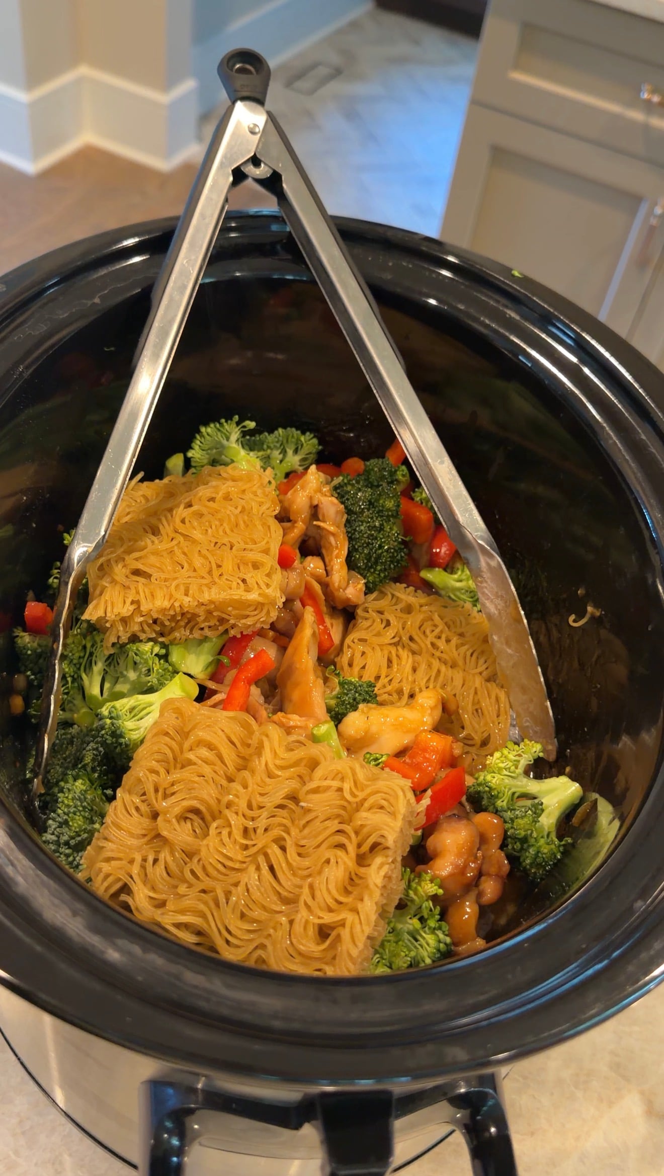 A slow cooker filled with uncooked ramen noodles, broccoli, red bell peppers, chicken, and sauce, with metal tongs resting inside.