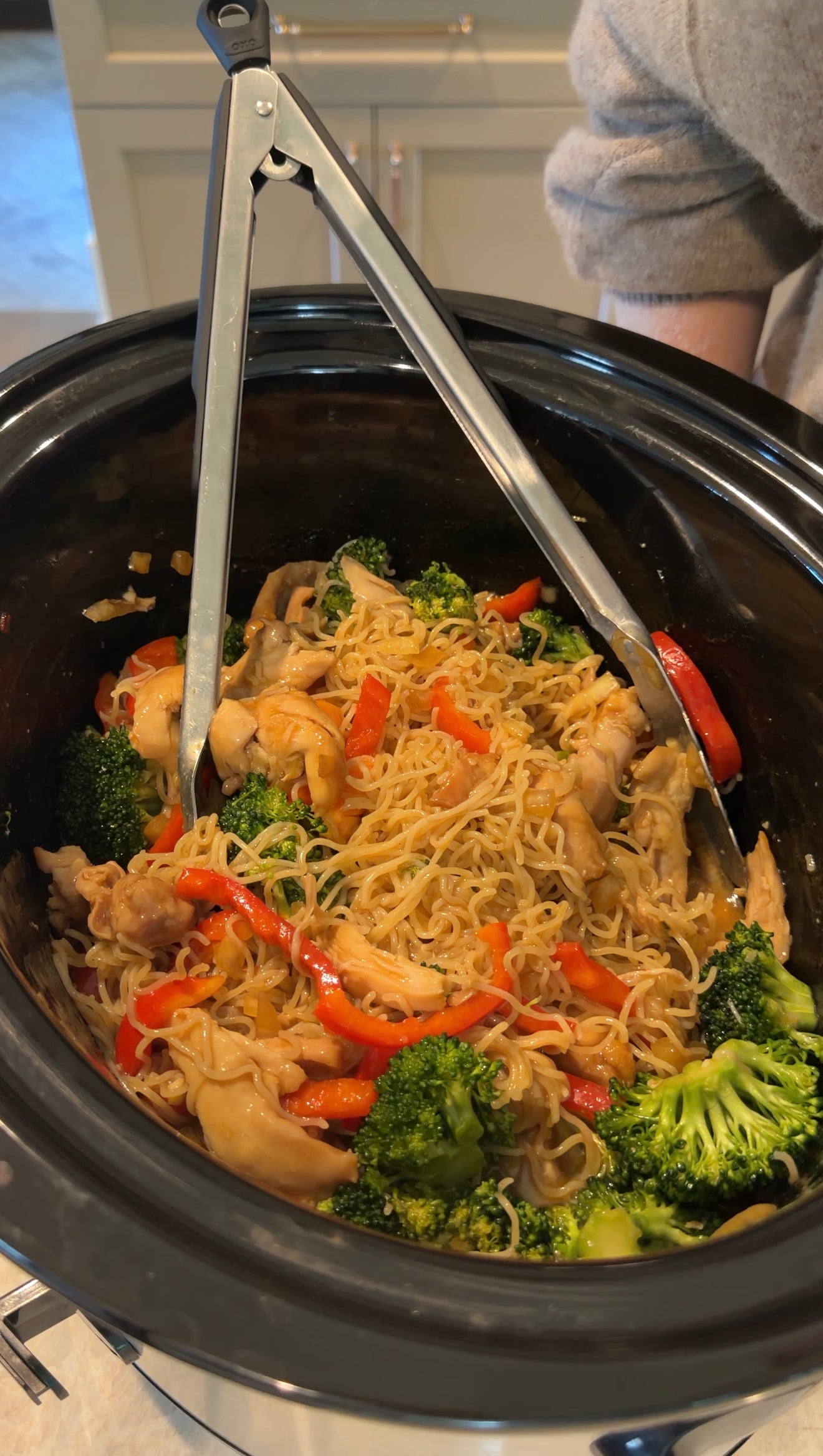 Noodles with chicken, broccoli, and red bell peppers in a slow cooker, with metal tongs resting on top, ready to serve.