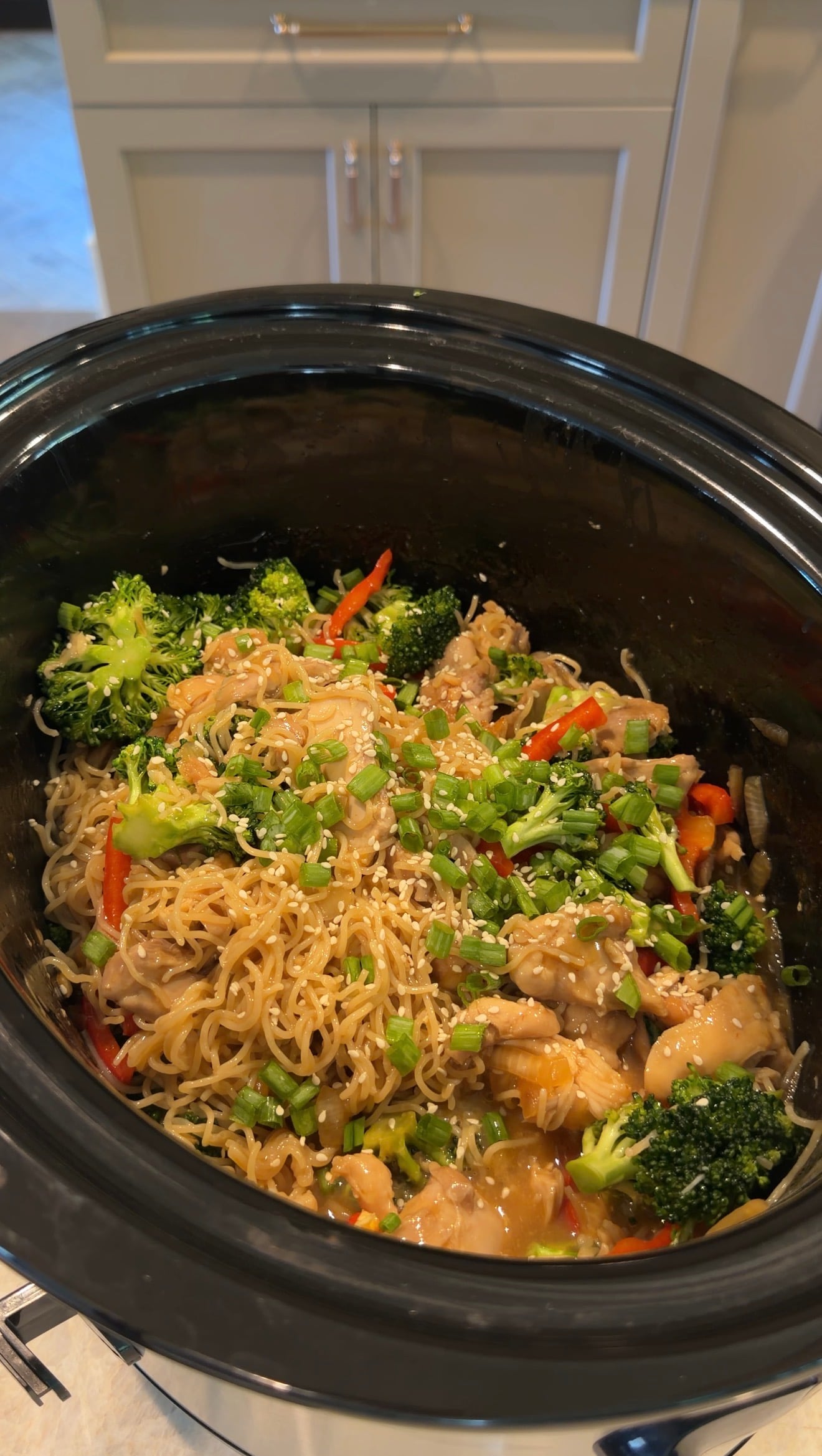 A crockpot filled with cooked noodles, broccoli, chicken, red bell peppers, chopped green onions, and sesame seeds in a savory sauce.