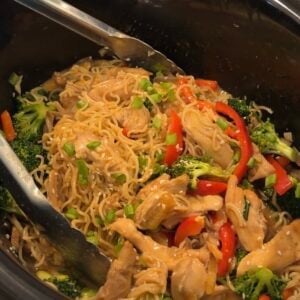 Chicken, noodles, broccoli, and red bell peppers in a slow cooker, mixed with sauce and garnished with chopped green onions. Metal tongs rest on the noodles, ready for serving.
