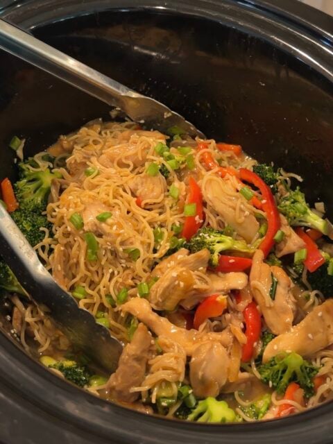Chicken, noodles, broccoli, and red bell peppers in a slow cooker, mixed with sauce and garnished with chopped green onions. Metal tongs rest on the noodles, ready for serving.