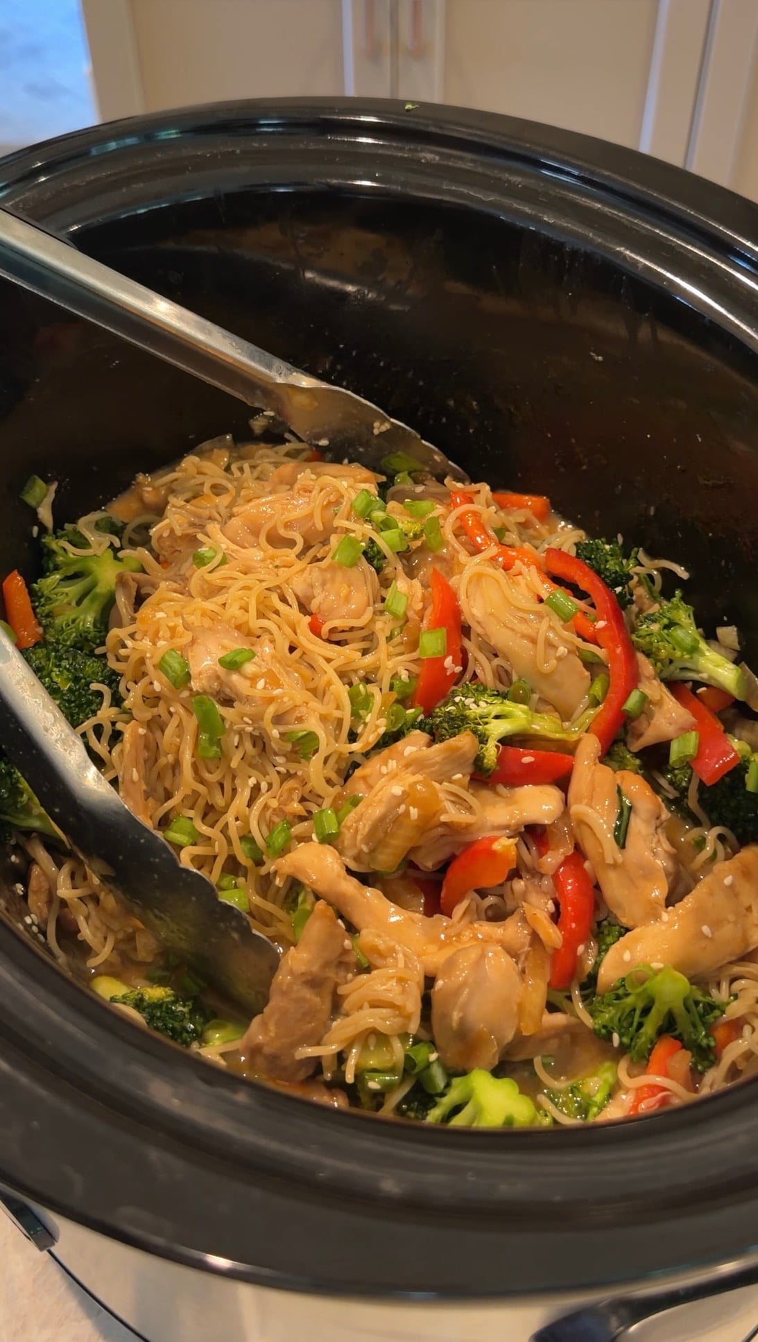 Chicken, noodles, broccoli, and red bell peppers in a slow cooker, mixed with sauce and garnished with chopped green onions. Metal tongs rest on the noodles, ready for serving.
