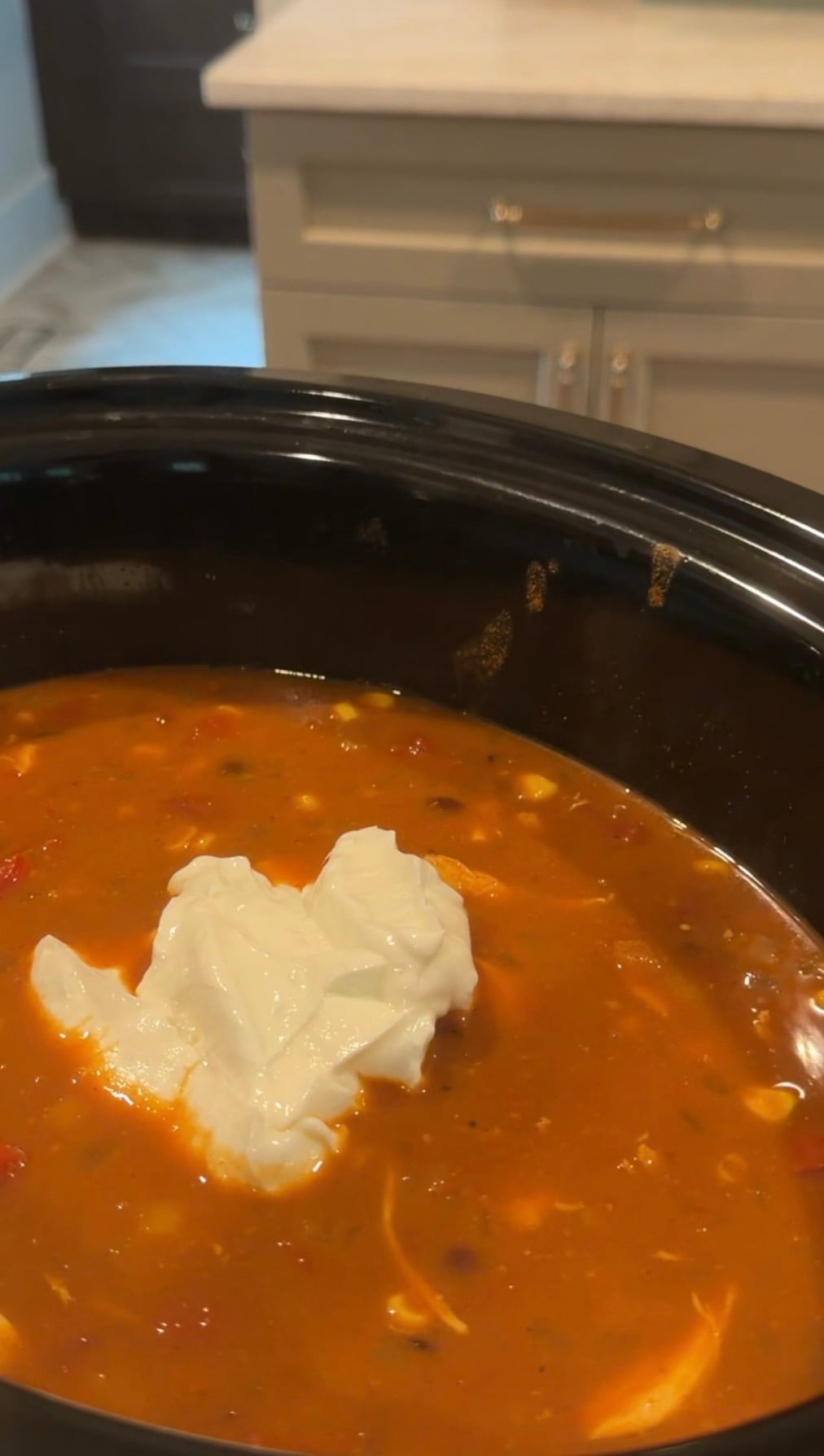 A close-up of a slow cooker filled with orange-red soup, topped with a dollop of sour cream. The soup contains visible corn and other vegetables. Kitchen cabinets are blurred in the background.