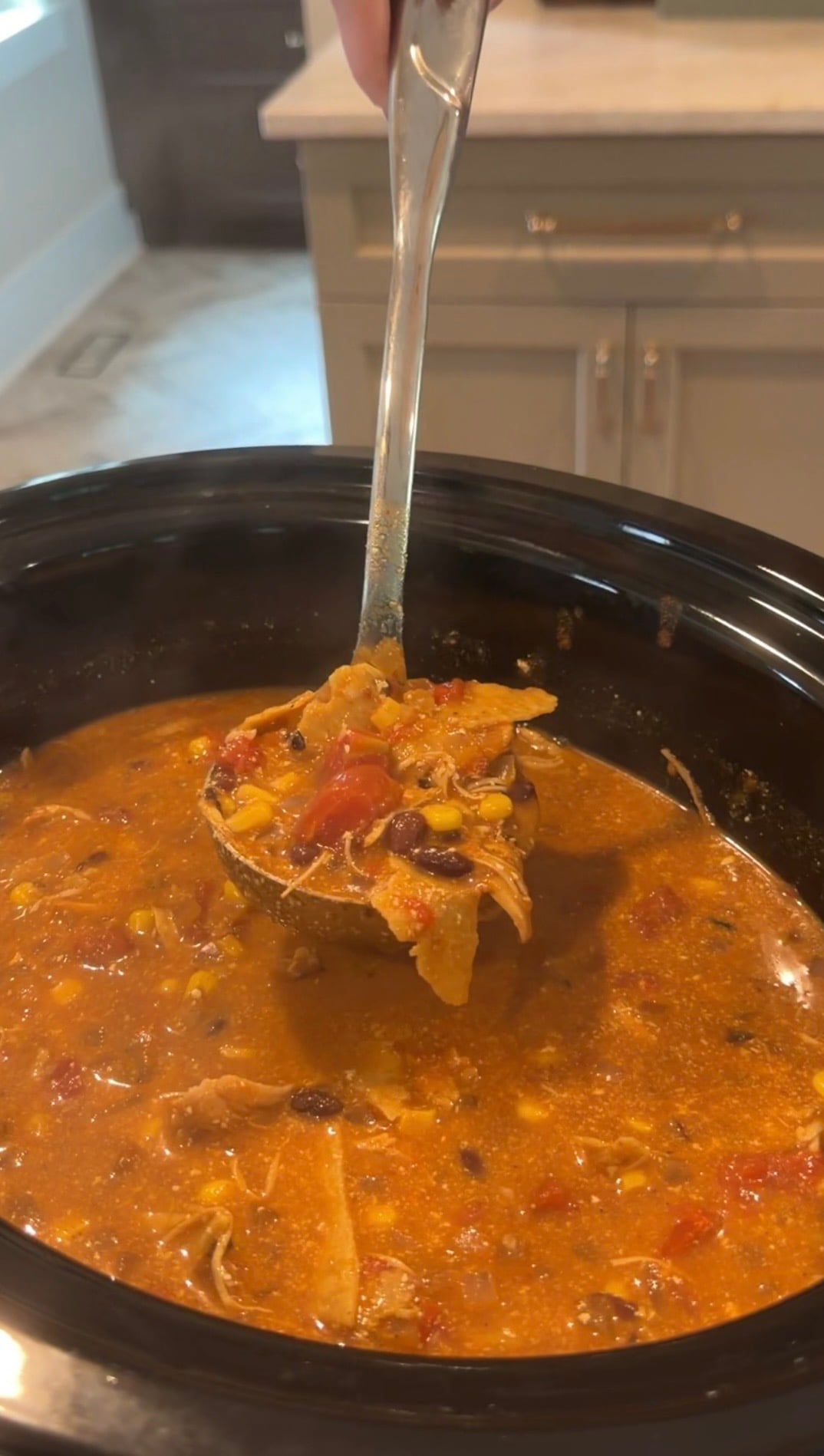 A ladle lifts a serving of hearty soup with beans, tomatoes, and shredded meat from a black slow cooker in a kitchen.