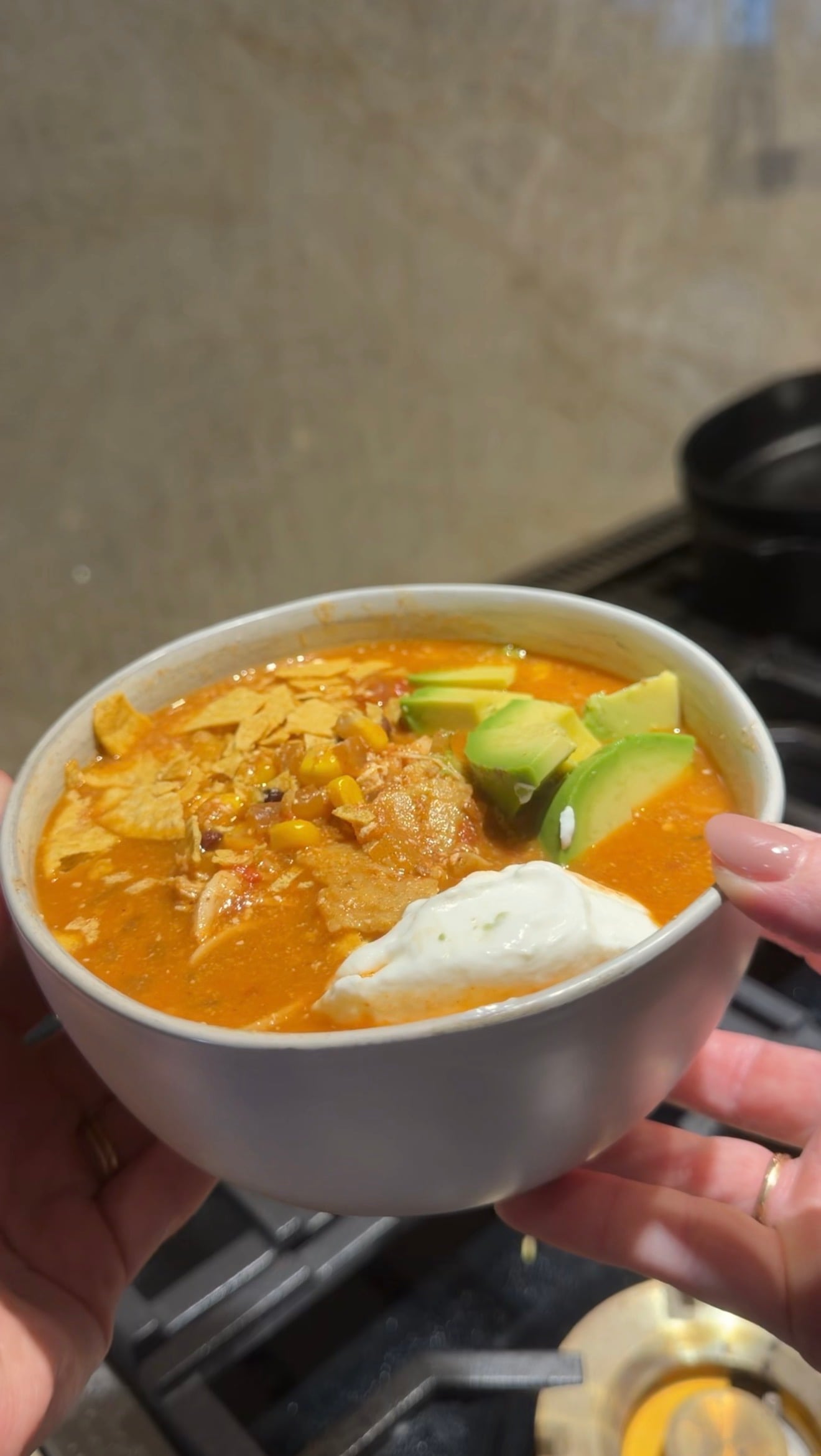 A hand holds a bowl of creamy orange soup topped with shredded chicken, avocado slices, tortilla strips, and a dollop of sour cream, with a stove visible in the background.