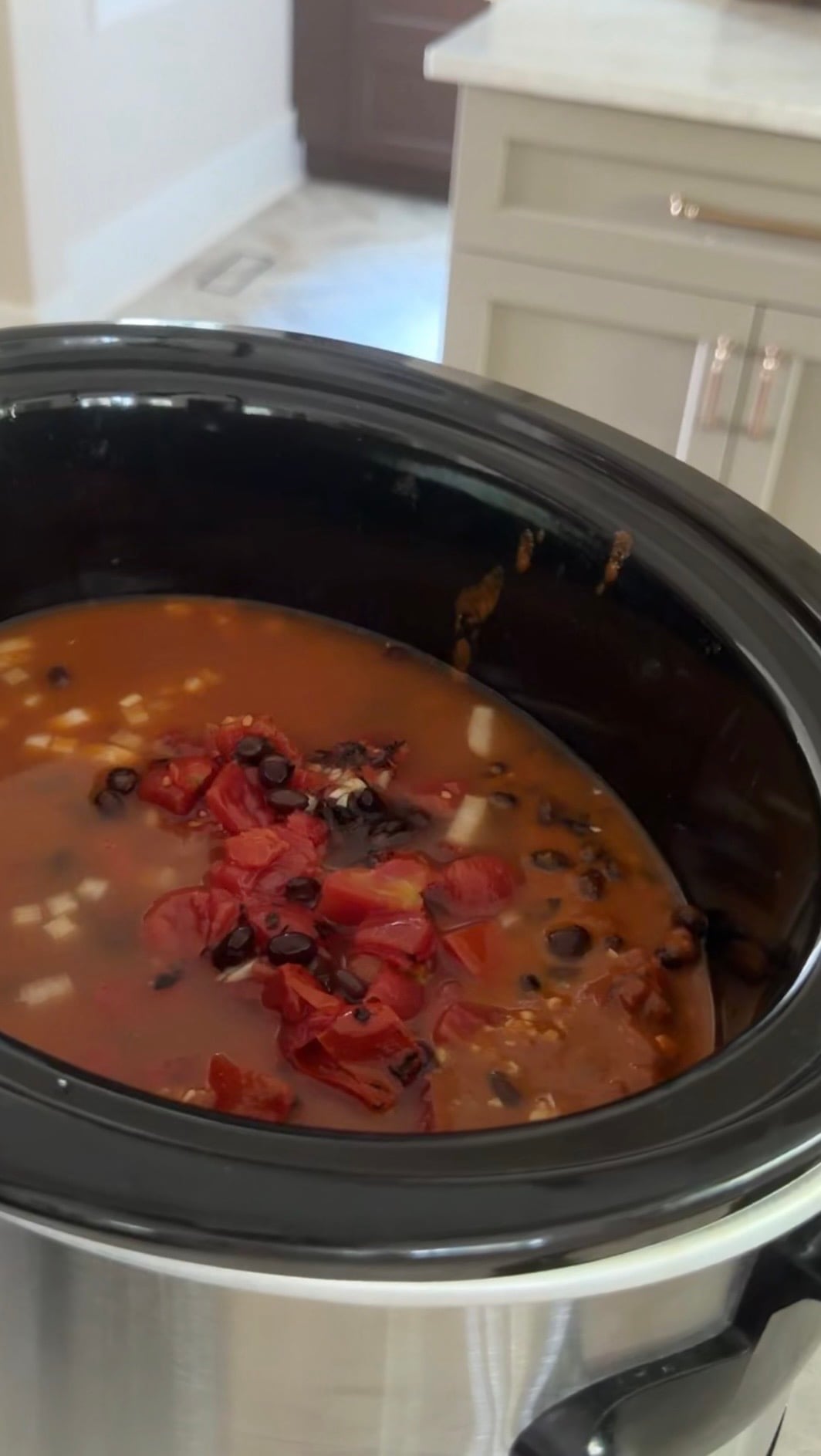 A slow cooker filled with a soup or stew containing black beans, chopped tomatoes, onions, and a red-orange broth sits on a kitchen counter.