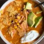 A bowl of tortilla soup topped with avocado slices, tortilla chips, corn, shredded chicken, and a dollop of sour cream, with a spoon resting on the bowl, set on a brown wooden surface.