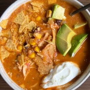 A bowl of tortilla soup topped with avocado slices, tortilla chips, corn, shredded chicken, and a dollop of sour cream, with a spoon resting on the bowl, set on a brown wooden surface.