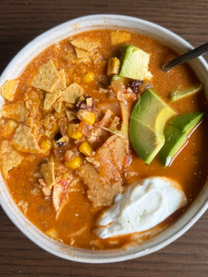 A bowl of tortilla soup topped with avocado slices, tortilla chips, corn, shredded chicken, and a dollop of sour cream, with a spoon resting on the bowl, set on a brown wooden surface.