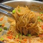 Tongs holding noodles mixed with vegetables like edamame and red peppers in a pan with sauce.