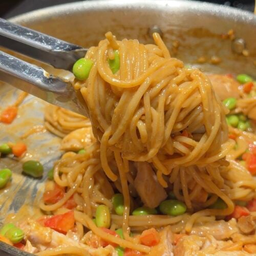 Tongs holding noodles mixed with vegetables like edamame and red peppers in a pan with sauce.