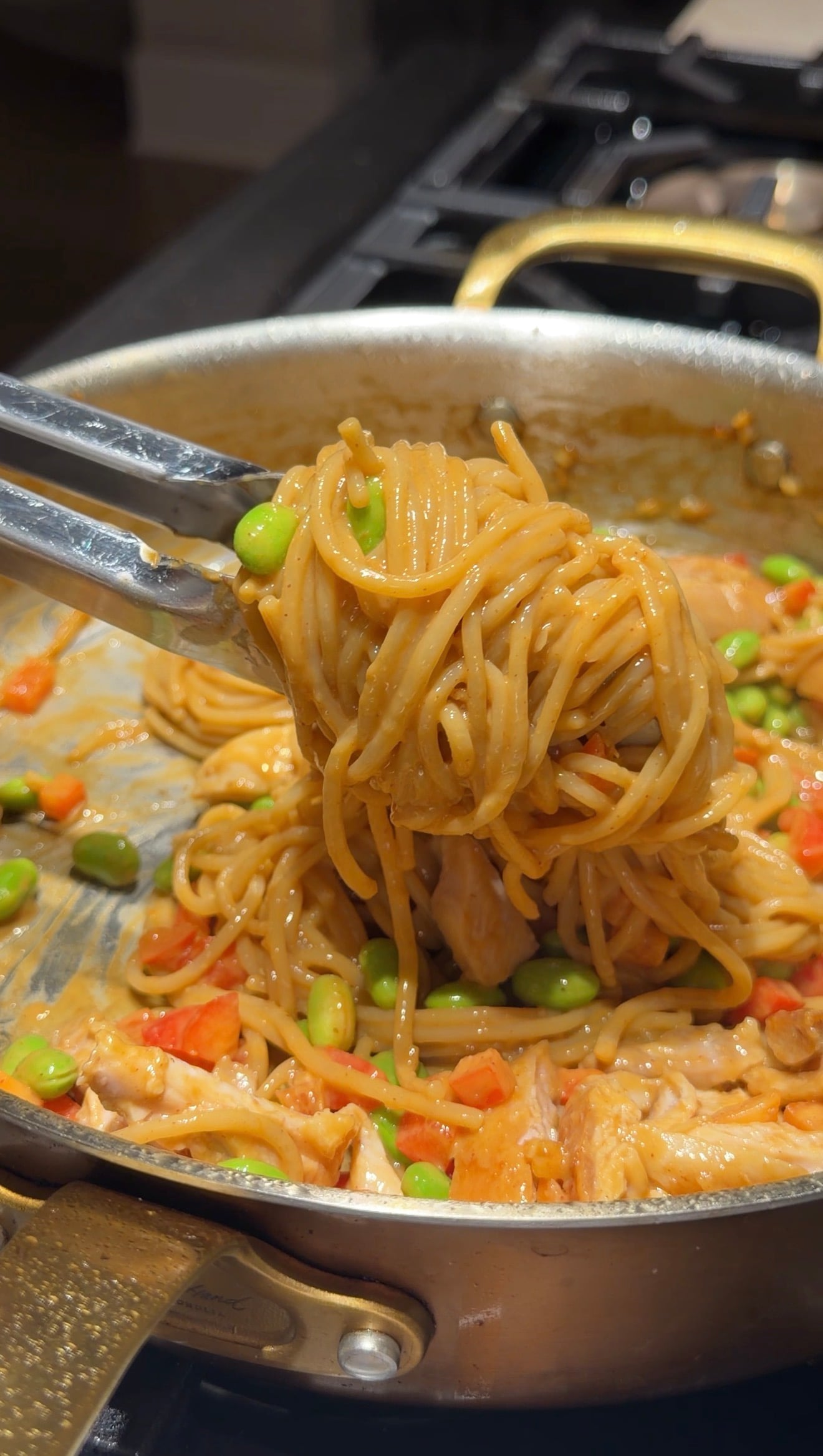 Tongs holding noodles mixed with vegetables like edamame and red peppers in a pan with sauce.