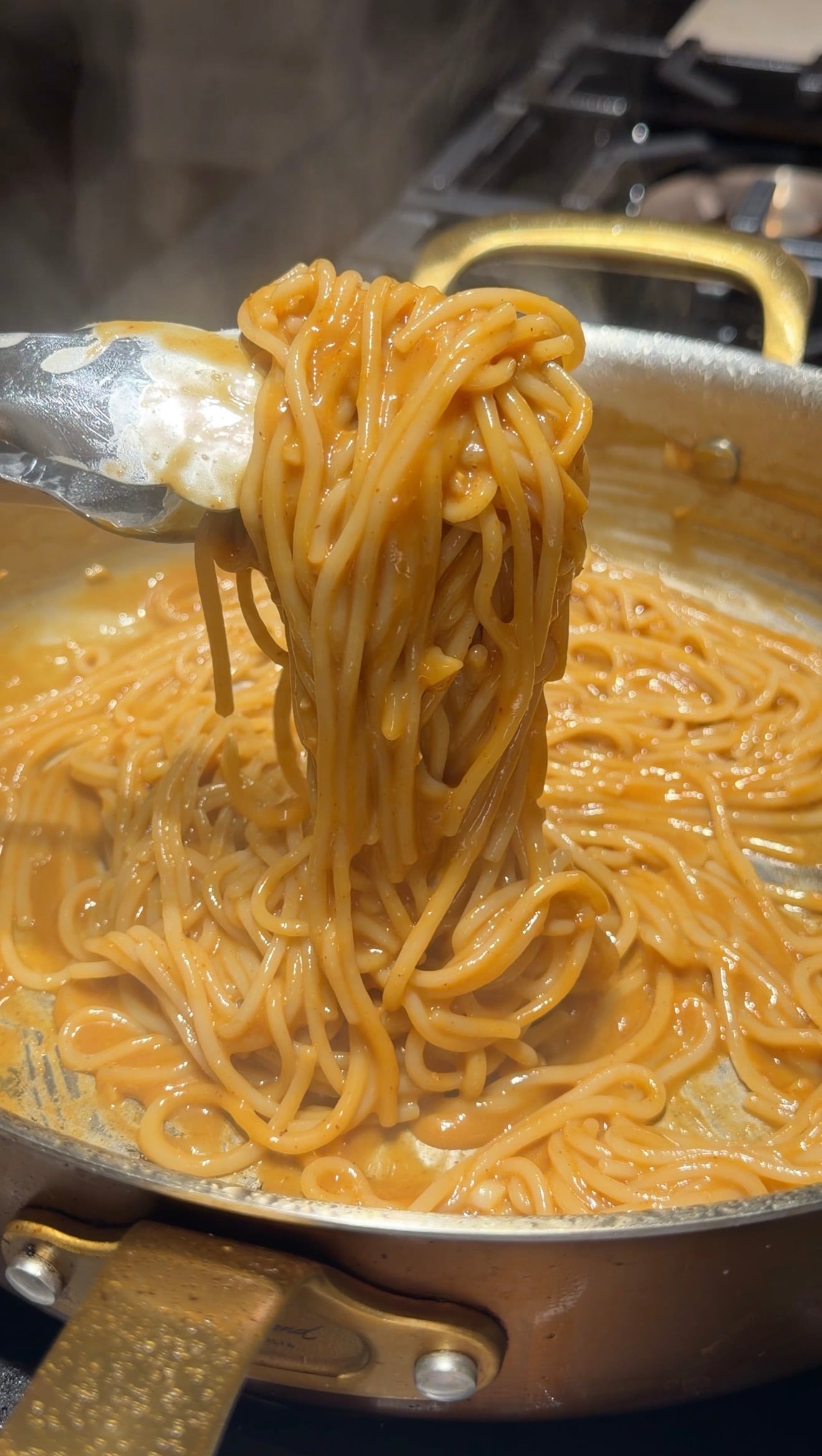 Tongs lift a serving of creamy, orange-colored spaghetti from a pan, showing a thick sauce coating the noodles.