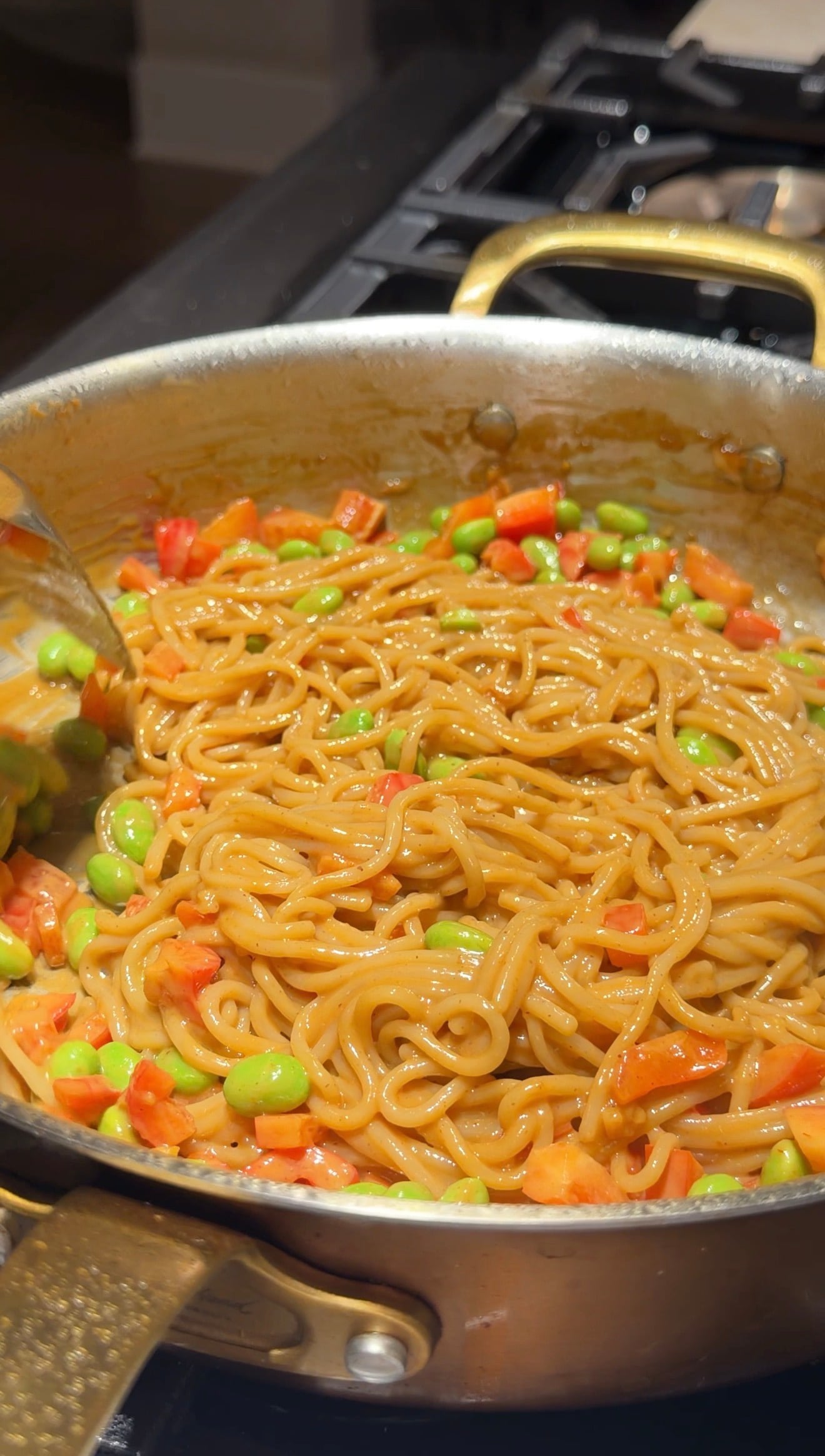 Cooked noodles in a large pan, mixed with diced carrots and green edamame beans, being stirred with a metal spoon. The noodles are coated in a glossy sauce.