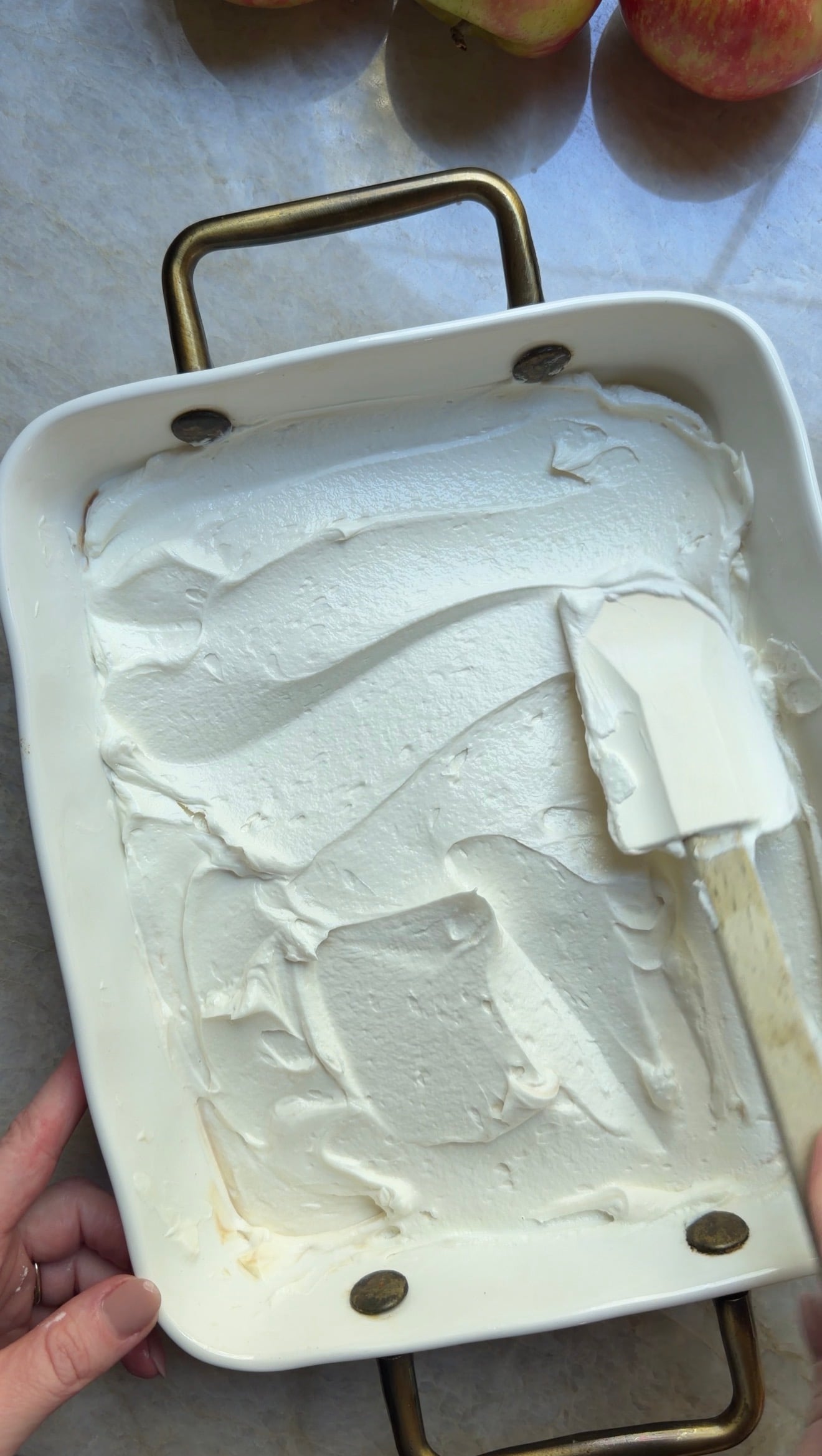 A hand spreads a thick layer of white whipped cream in a rectangular baking dish using a spatula. Apples are partially visible at the top of the image.