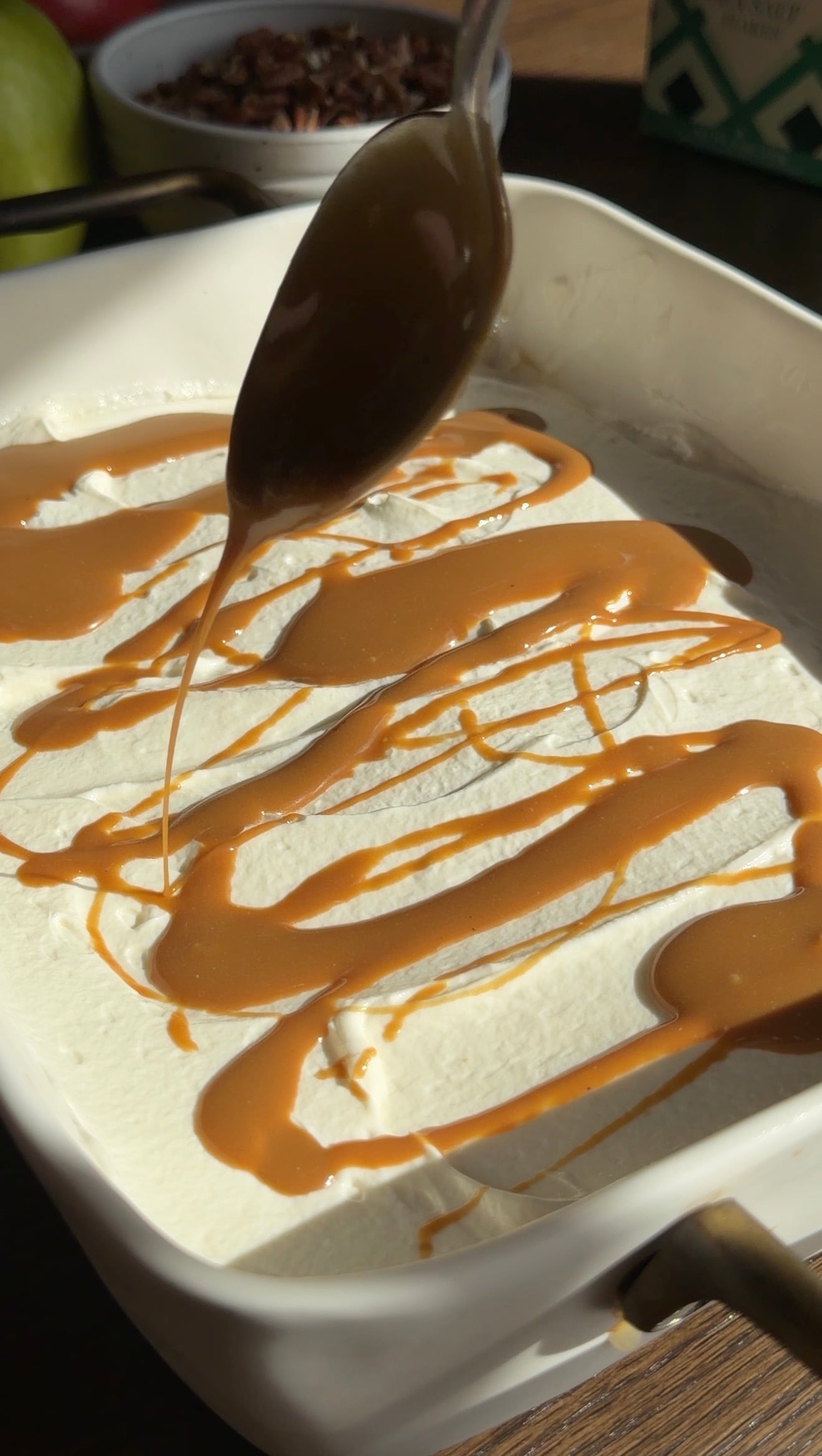 A spoon drizzles caramel sauce over a layer of whipped cream in a white dish, creating swirls and lines across the creamy surface.