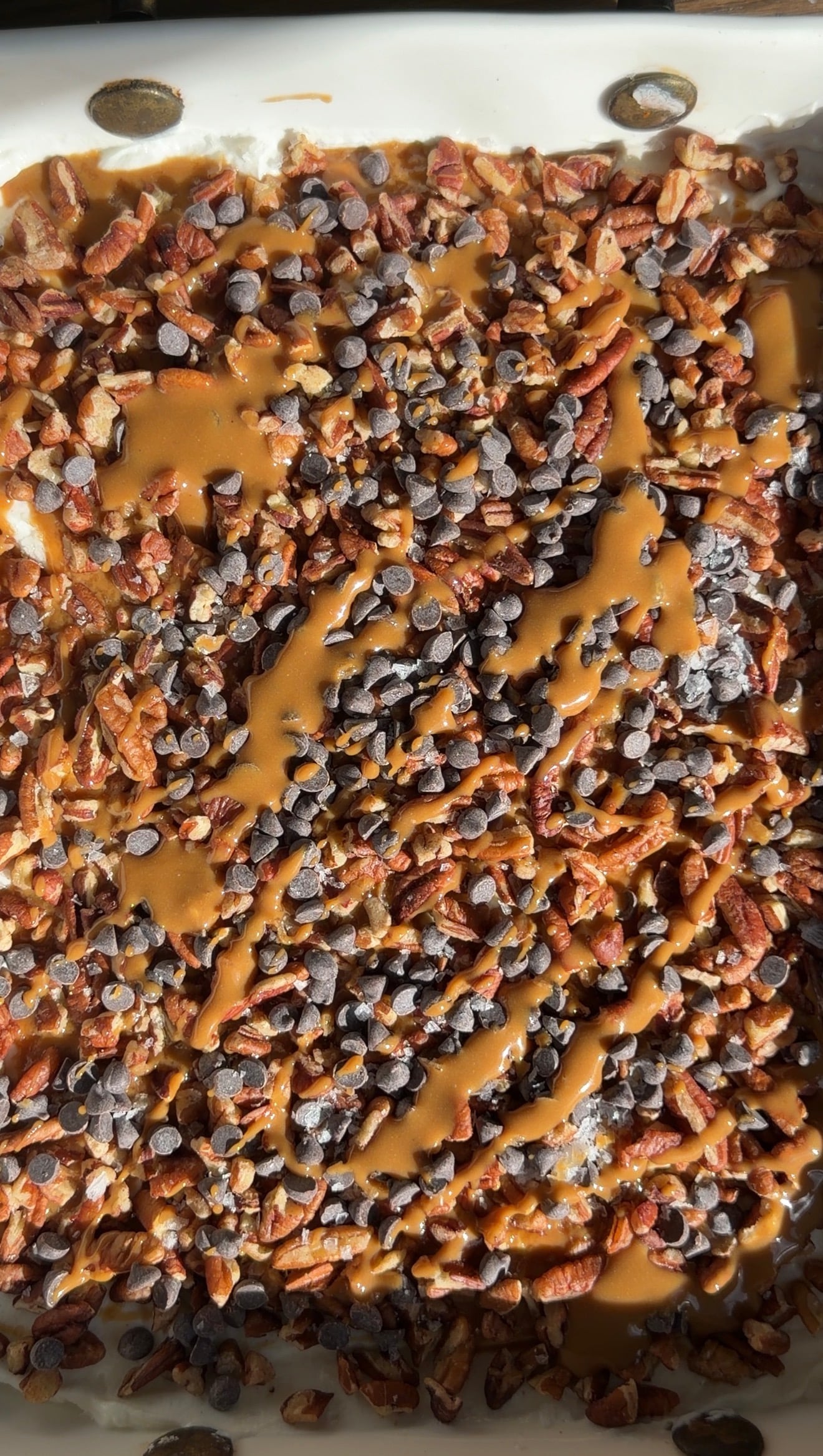 A close-up of a dessert topped with pecans, mini chocolate chips, and a drizzle of caramel sauce, all in a white baking dish. Sunlight highlights the textures and rich colors.