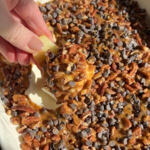 A hand dips an apple slice into a dish filled with caramel, chopped pecans, and mini chocolate chips.