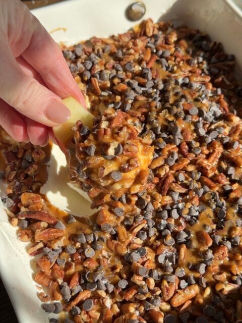 A hand dips an apple slice into a dish filled with caramel, chopped pecans, and mini chocolate chips.