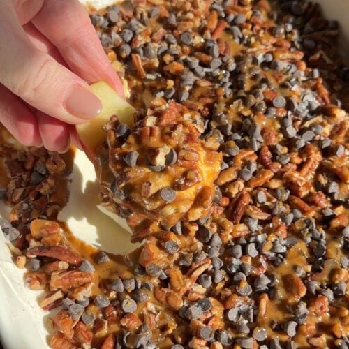 A hand dips an apple slice into a dish filled with caramel, chopped pecans, and mini chocolate chips.
