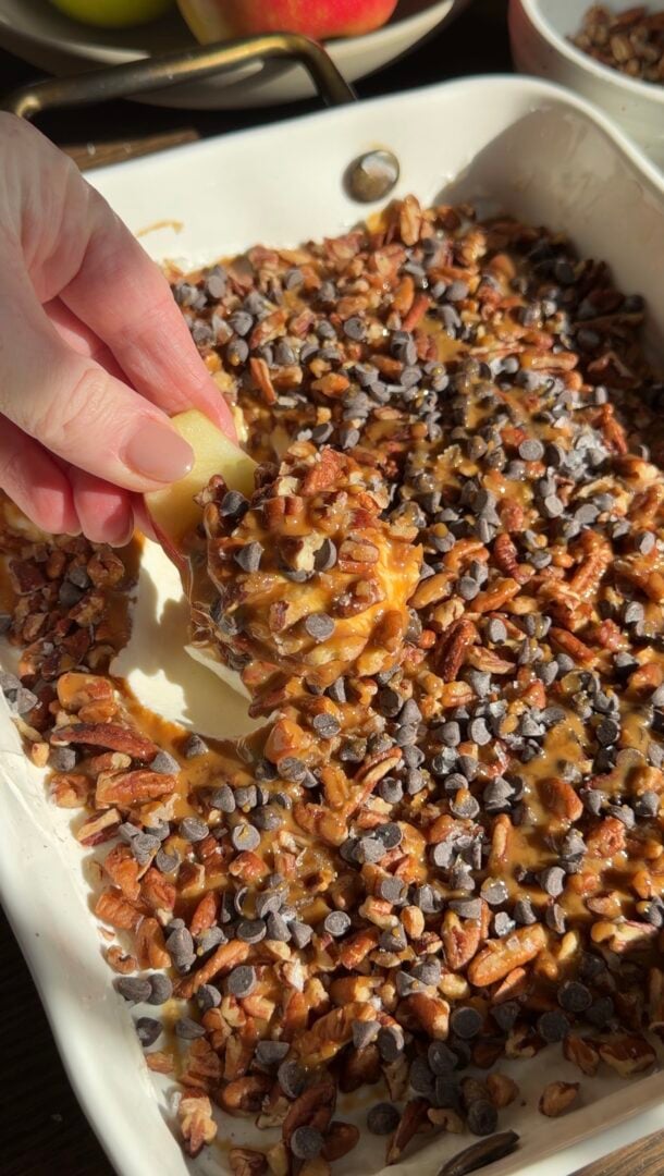 A hand dips an apple slice into a dish filled with caramel, chopped pecans, and mini chocolate chips.
