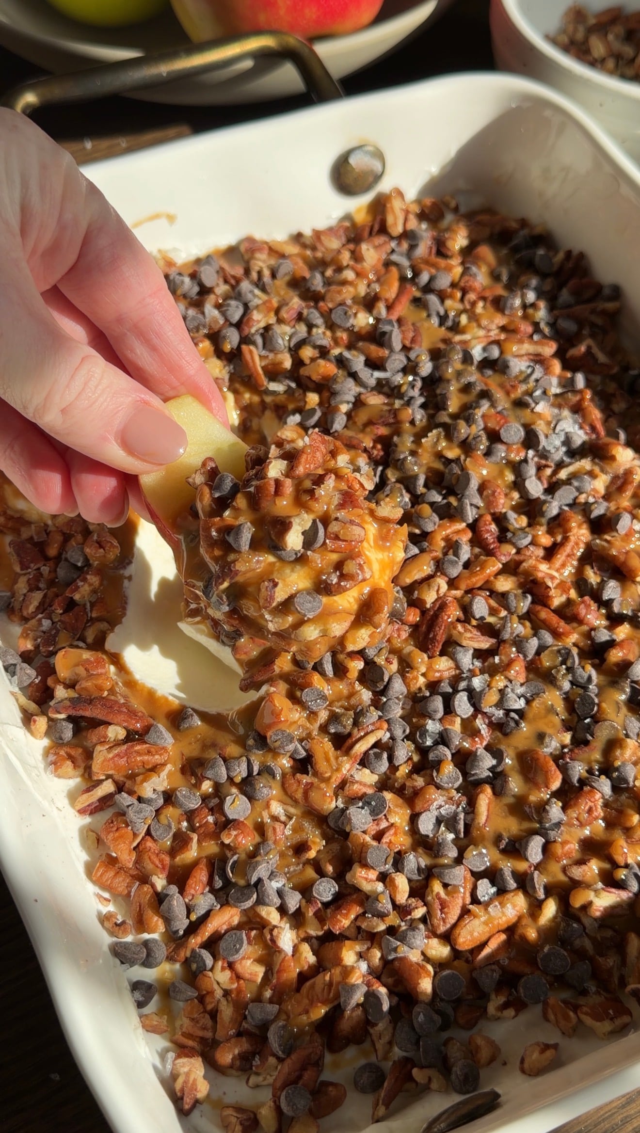 A hand dips an apple slice into a dish filled with caramel, chopped pecans, and mini chocolate chips.