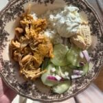 A decorative plate with shredded chicken, white rice, cucumber salad with red onion, a dollop of tzatziki, and hummus. Sunlight shines on the plate, which is held by a person wearing light-colored clothing.
