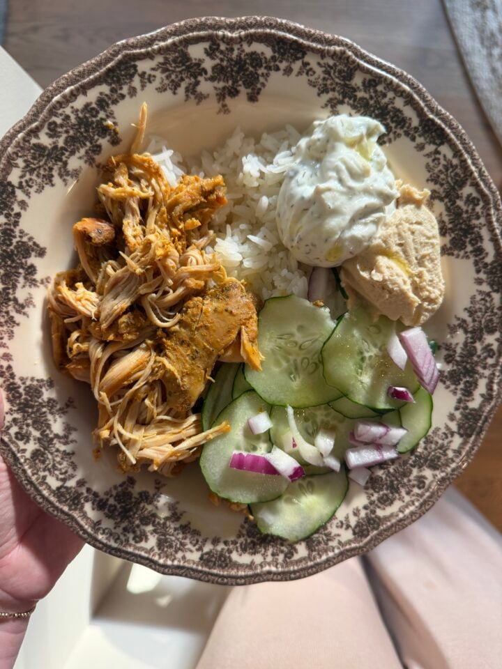 A decorative plate with shredded chicken, white rice, cucumber salad with red onion, a dollop of tzatziki, and hummus. Sunlight shines on the plate, which is held by a person wearing light-colored clothing.