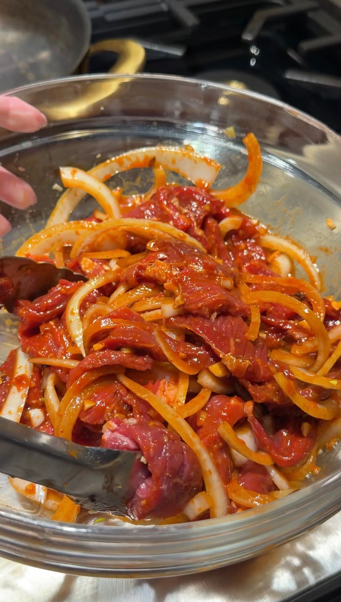 A glass bowl filled with raw beef strips, sliced onions, and red marinade being mixed with tongs, ready for cooking.