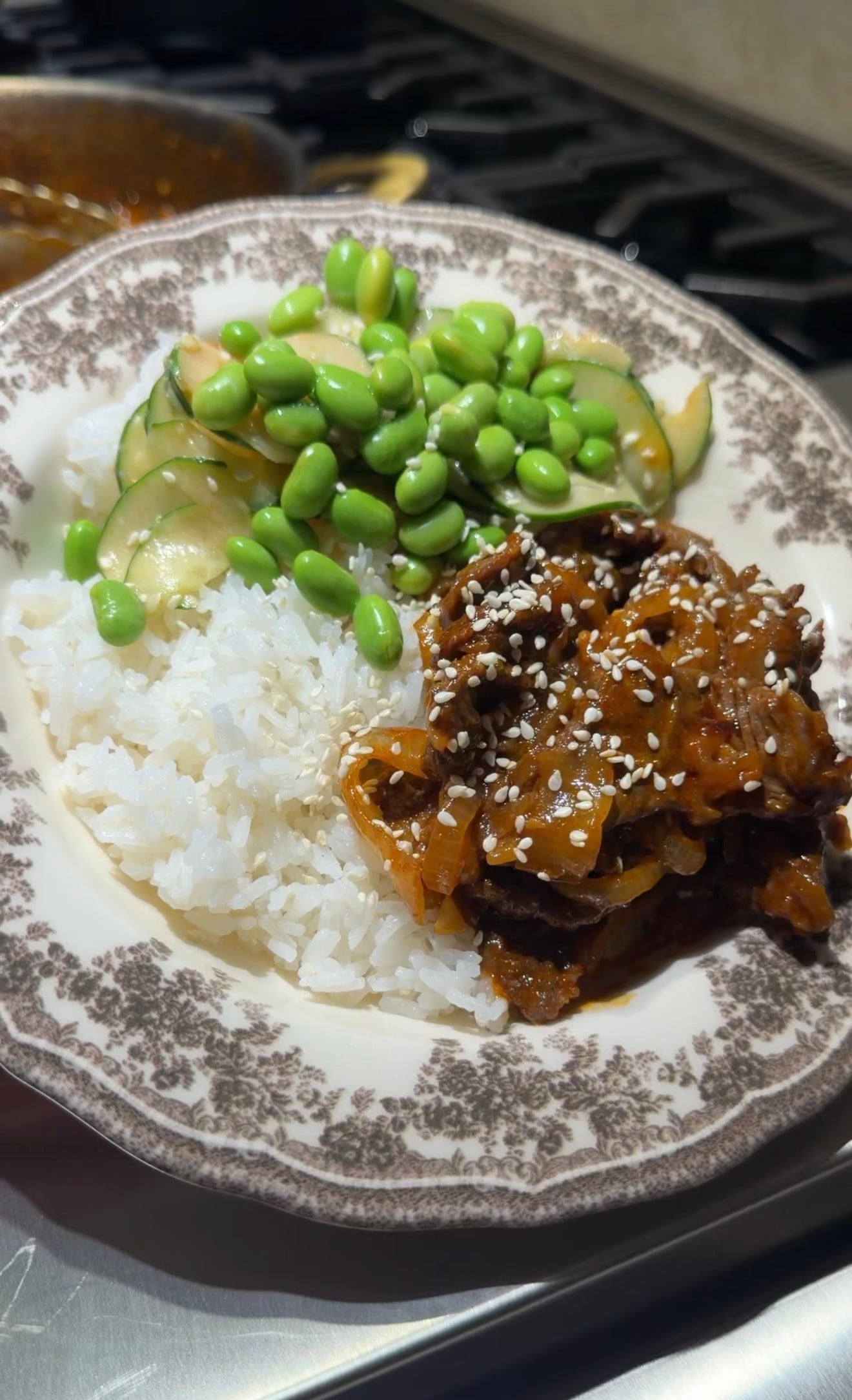 A plate with white rice, cucumber and edamame salad, and a portion of saucy beef topped with sesame seeds. The food is served on a floral-patterned plate.