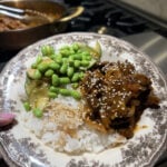 A plate of white rice topped with glazed mushrooms, sesame seeds, sliced cucumbers, and edamame, held in front of a stove with a pan in the background.