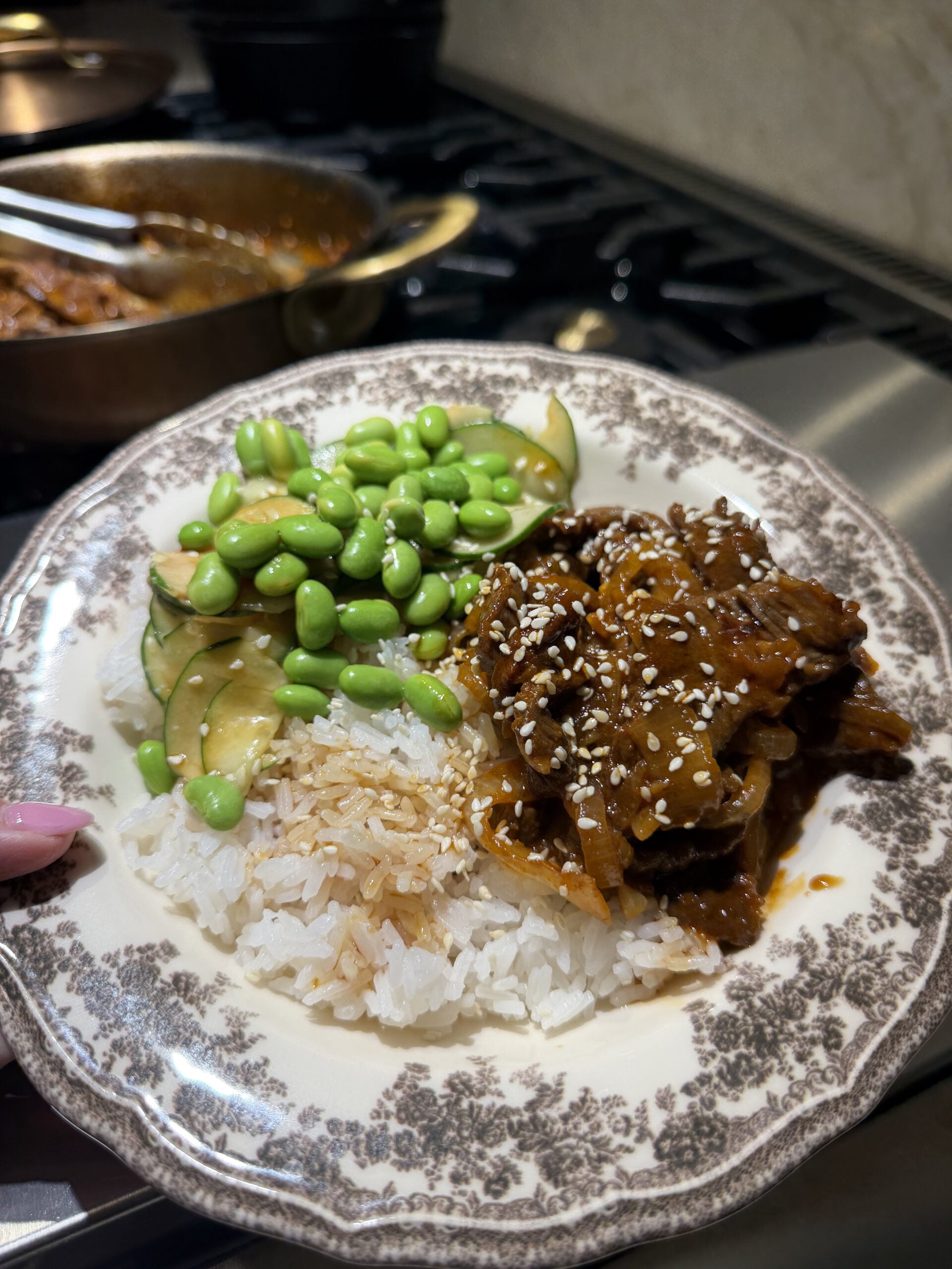 A plate of white rice topped with glazed mushrooms, sesame seeds, sliced cucumbers, and edamame, held in front of a stove with a pan in the background.