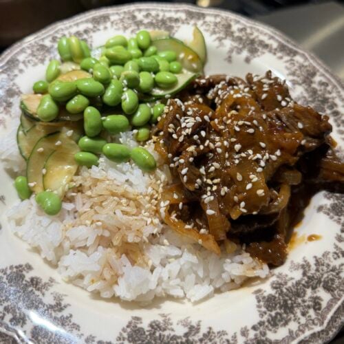 A plate of white rice topped with sliced cucumbers, edamame, and saucy beef with sesame seeds, served on a decorative dish—these Korean beef bowls bring together fresh flavors and vibrant textures for a satisfying meal.