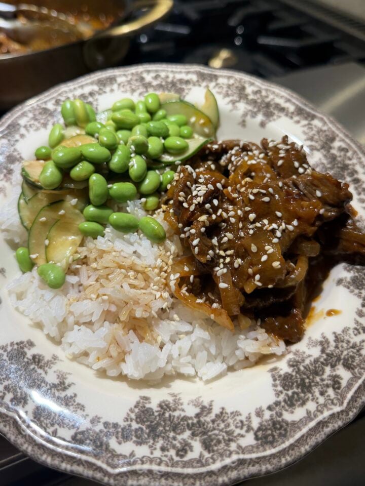 A plate of white rice topped with sliced cucumbers, edamame, and saucy beef with sesame seeds, served on a decorative dish—these Korean beef bowls bring together fresh flavors and vibrant textures for a satisfying meal.