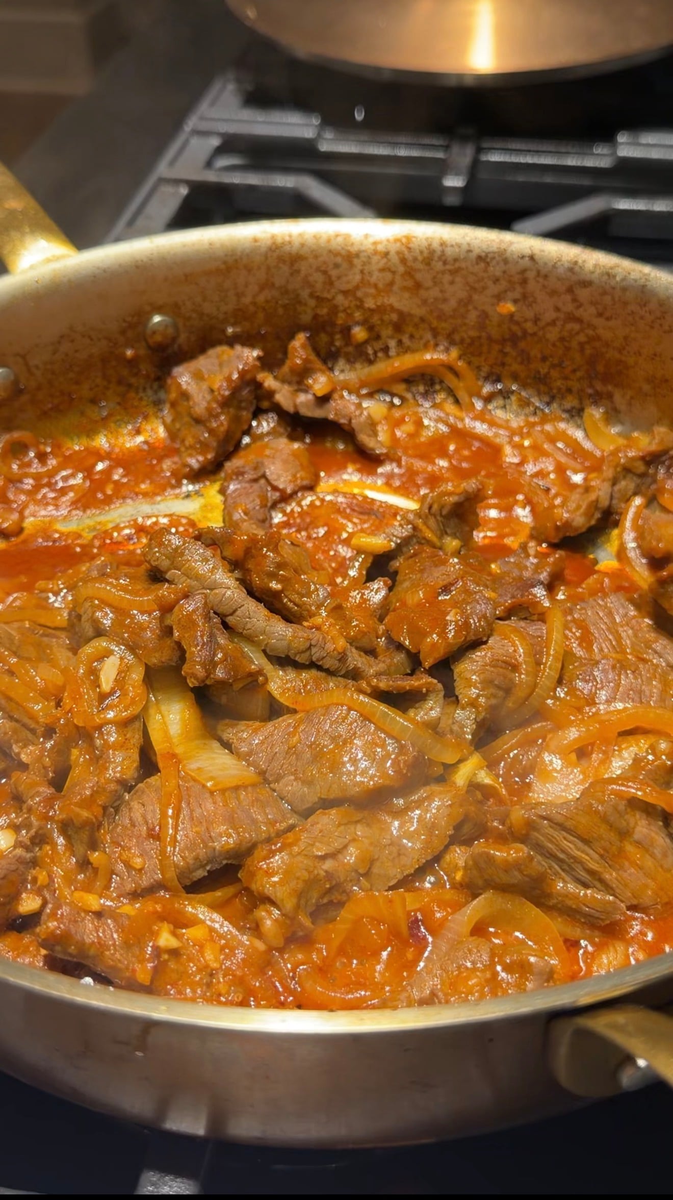 Strips of beef and onions are simmering in a rich, red tomato-based sauce inside a large, shallow pan on a stovetop.
