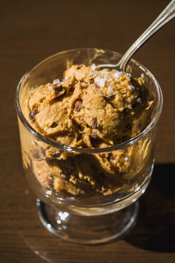 A glass cup filled with scoops of brown ice cream or cookie dough, topped with sea salt flakes and chocolate chips, with a spoon inside.