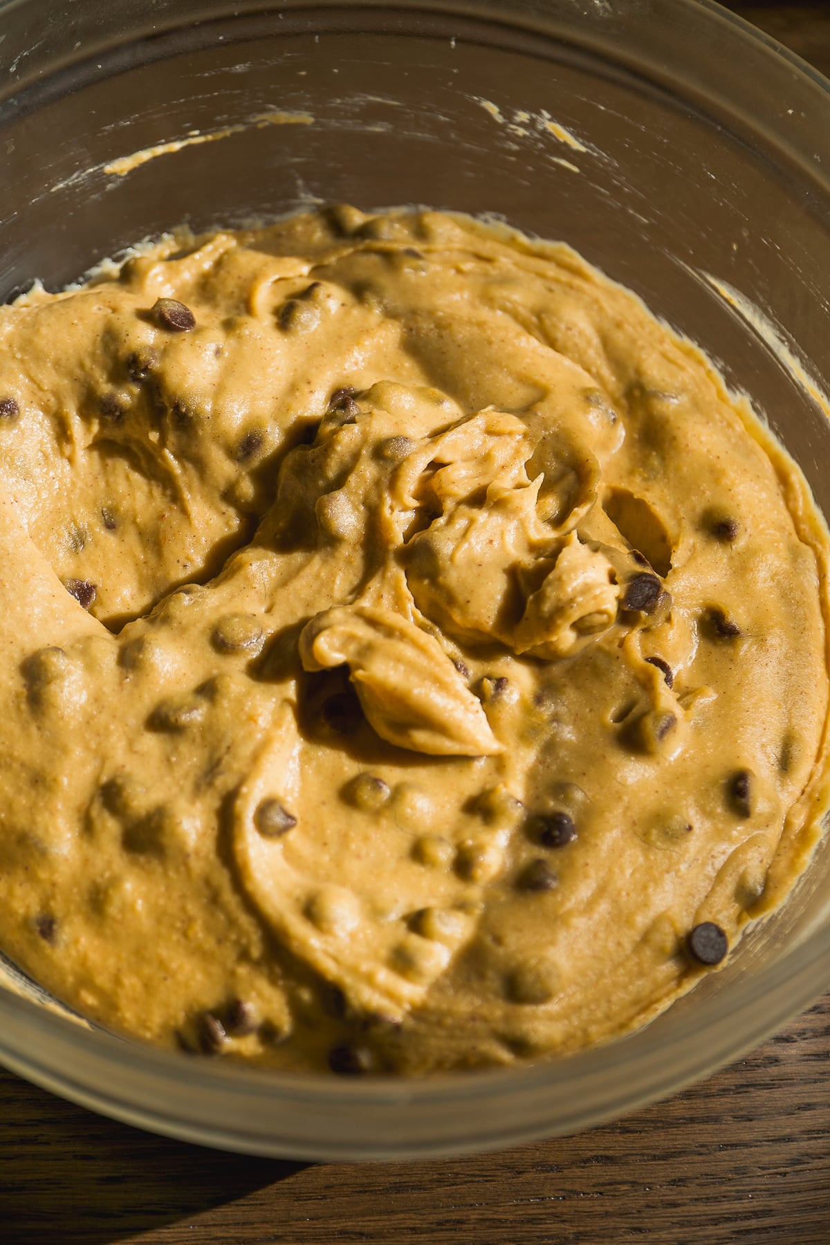 A clear glass bowl filled with thick cookie dough mixed with chocolate chips, sitting on a wooden surface in natural sunlight.