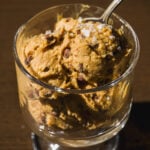 A glass cup filled with scoops of chocolate chip ice cream topped with coarse sea salt, with a spoon sticking out, on a dark wooden surface.