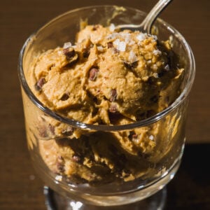 A glass cup filled with scoops of chocolate chip ice cream topped with coarse sea salt, with a spoon sticking out, on a dark wooden surface.