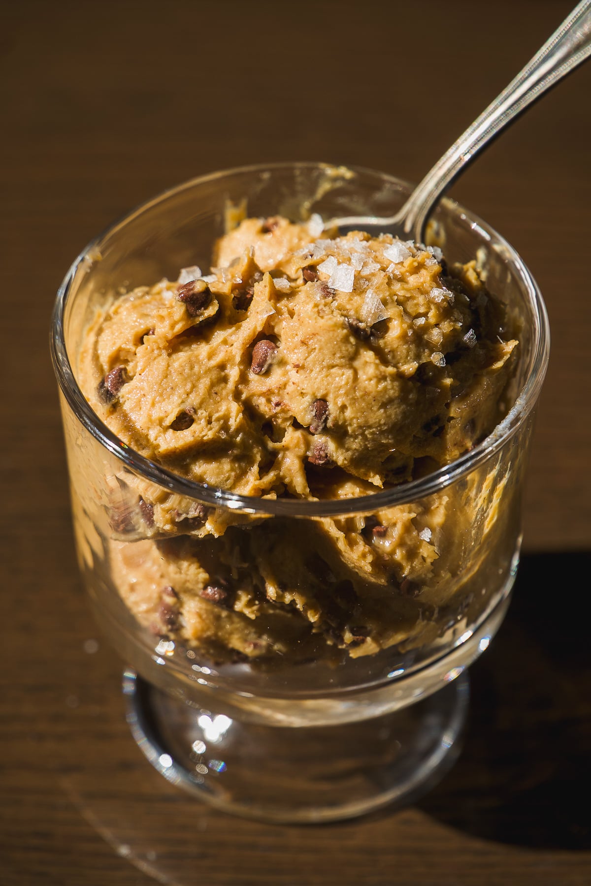 A glass cup filled with scoops of chocolate chip ice cream topped with coarse sea salt, with a spoon sticking out, on a dark wooden surface.