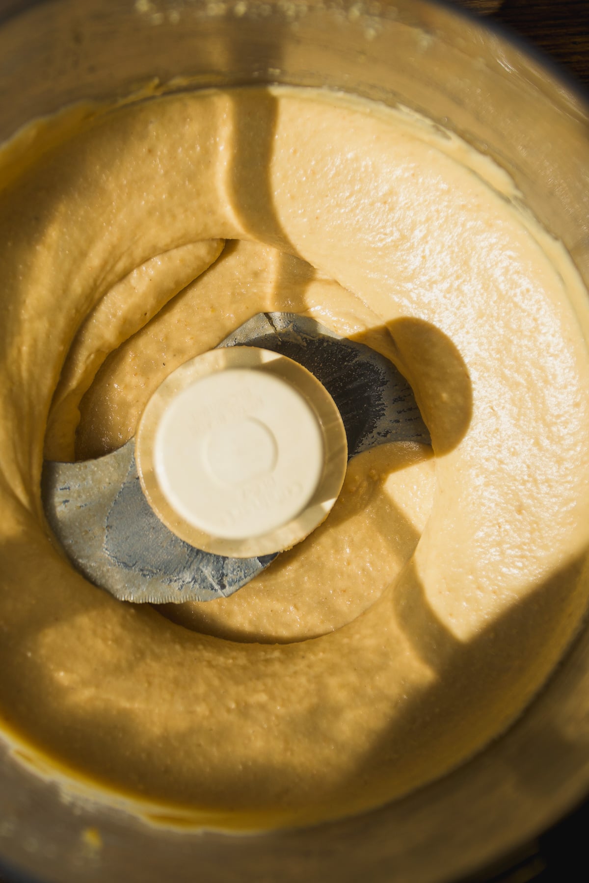 Creamy, light brown mixture being blended in a food processor, with visible metal blade and smooth texture forming a swirl pattern under natural light.